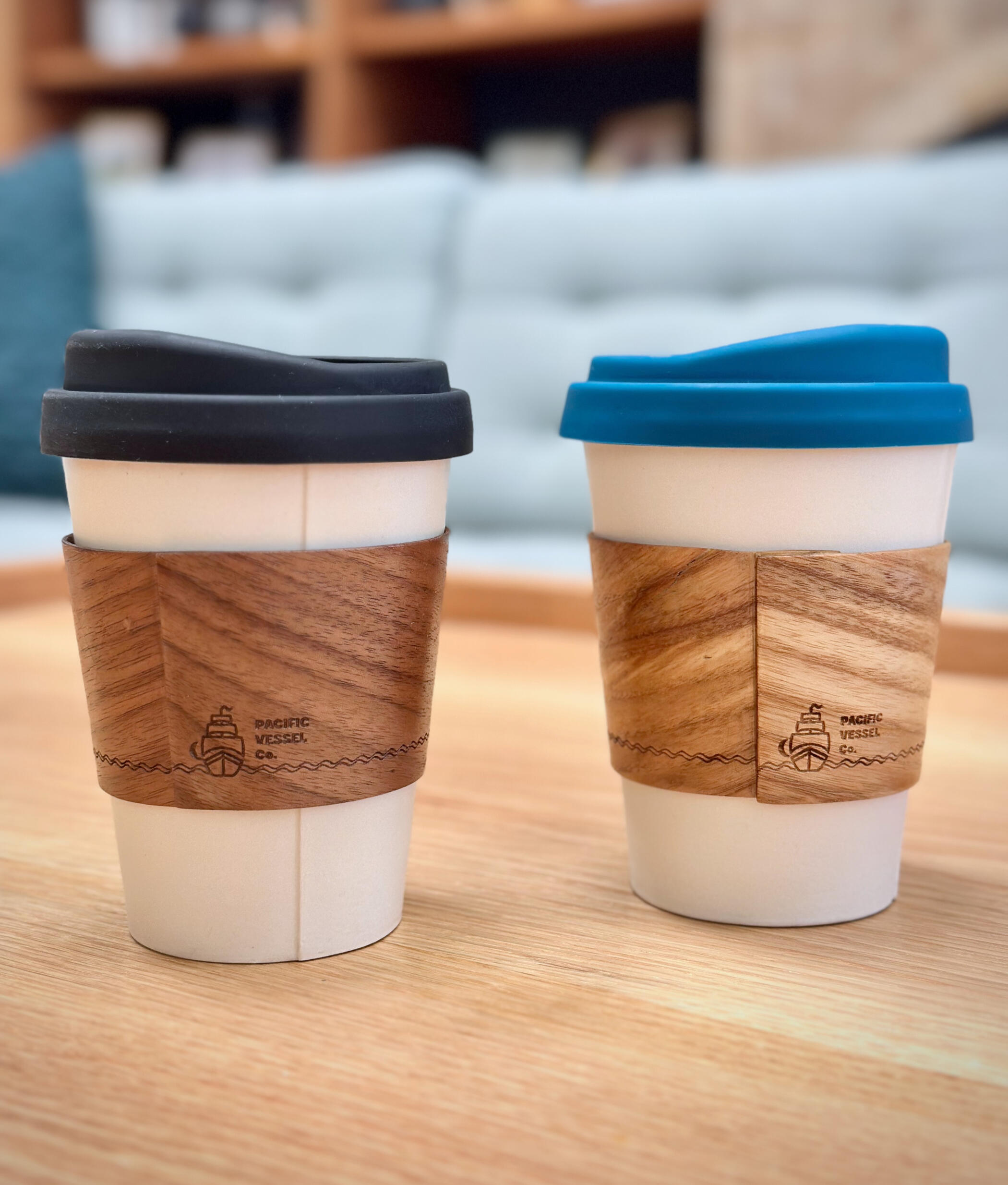 Two ceramic coffee mugs in the style of paper takeout mugs, with wooden sleeves and silicon lids, sitting on top of a wooden table